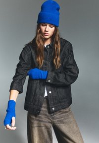Giacca di denim nera con bottoni argentati, abbinata a un berretto di lana blu e guanti senza dita. Modello che indossa pantaloni beige larghi.