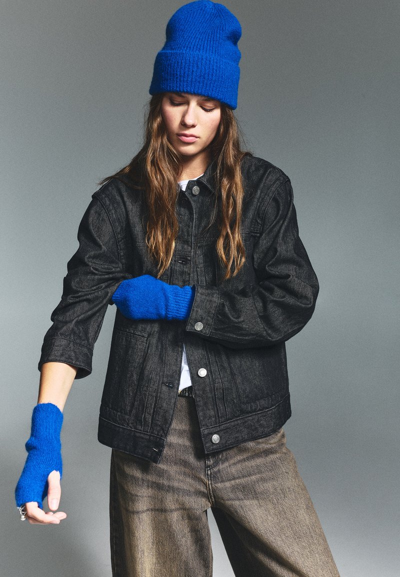 Giacca di denim nera con bottoni argentati, abbinata a un berretto di lana blu e guanti senza dita. Modello che indossa pantaloni beige larghi.