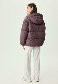 Lila Daunenjacke mit Kapuze, mit horizontalen Steppsektionen. Kombiniert mit grauen Jogginghosen und Sneakers. Minimalistisches Design.