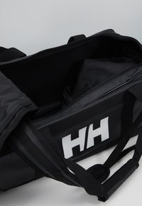 Bolsa de deporte negra hecha de tela duradera, con una gran abertura, cremallera negra y un destacado logotipo blanco. Se incluyen correas ajustables.