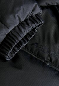 Chaqueta aislante negra con un tejido texturizado, puños fruncidos y un logo de marca visible. Diseño detallado con acentos minimalistas.