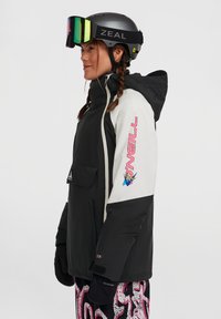 Zwart-witte skijas met een voorzak, voorzien van een kleurrijk logo op de mouw. Het model draagt een zwarte helm met spiegelende skibril.