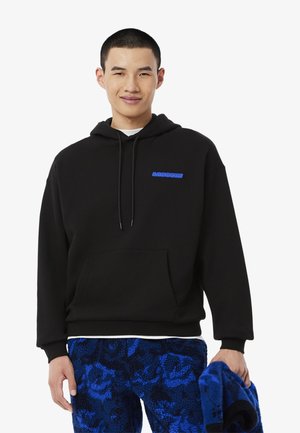 Haut à capuche noir avec une poche avant et un logo bleu. Modèle portant un pantalon texturé bleu, tenant un tissu bleu. Design simple et décontracté.
