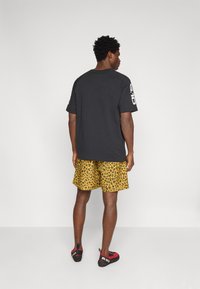 Black T-shirt with short sleeves; paired with yellow leopard print shorts. Black climbing shoes with red accents. Plain background.