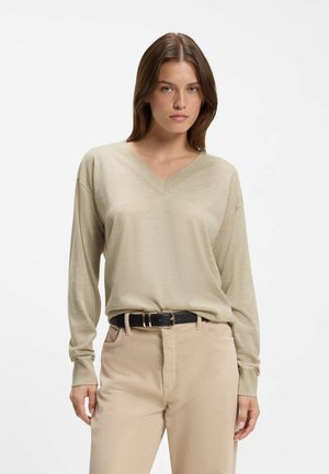 Kvinde iført en beige langærmet V-hals sweater, lysebeige bukser og et sort bælte, stående foran en ensfarvet hvid baggrund.