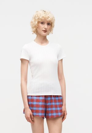 Νεαρό άτομο με καστανά ξανθά μπούκλες, που φοράει λευκή ριπ κοντομάνικη μπλούζα και κόκκινο-μπλε καρό σορτς, στέκεται μπροστά σε ένα απλό φόντο.