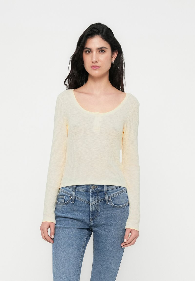 Μακρυμάνικο τοπ σε ανοιχτό κίτρινο χρώμα με λαιμόκοψη scoop και υφασμάτινη υφή, συνδυασμένο με μπλε τζιν παντελόνι. Απλός σχεδιασμός, εφαρμοστή σιλουέτα.