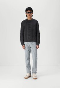 Pulover gri închis, împletit, cu guler rotund, asortat cu pantaloni albaștri deschiși și adidași bej. Accesoriile includ ochelari de soare negri.