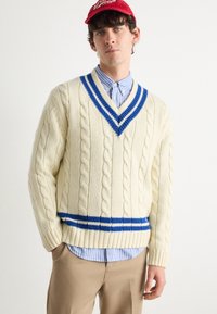 Maglione beige a treccia con scollatura a V profonda, dettagli a righe blu e vestibilità ampia, indossato sopra una camicia a righe blu chiaro.