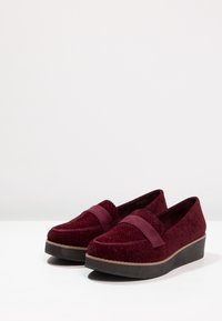 Chaussures bordeaux avec une tige texturée, une large sangle sur l'empeigne et une semelle épaisse et sombre. Tissu lisse et détails subtils.