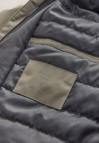 Vue intérieure d'une veste matelassée montrant une étiquette en tissu beige avec l'inscription "NEXT", la taille "M" et "Fabriqué en Chine".