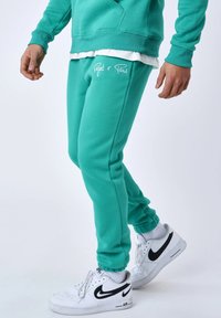 Pantalones de chándal turquesa con puños elásticos, que presentan un texto bordado en blanco "Pajet x Paris". Combinados con zapatillas blancas de Nike y calcetines blancos.