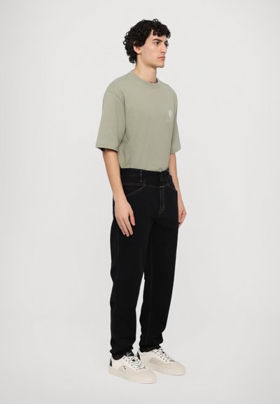 Camiseta de gran tamaño verde claro, de mangas cortas, con cuello redondo. Pantalones negros con costuras prominentes, combinados con zapatillas blancas. Diseño simple.