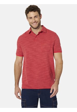Lächelnder Mann mit rotem kurzärmligem Poloshirt und dunkelblauer Cargohose, der vor einem schlichten hellen Hintergrund steht.