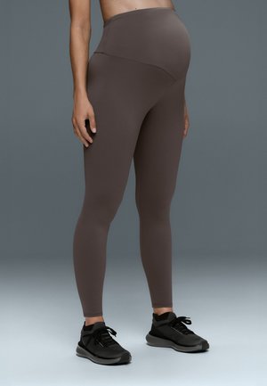 Personne enceinte portant des leggings de maternité marron foncé et des chaussures de sport noires, debout sur un fond gris uni.