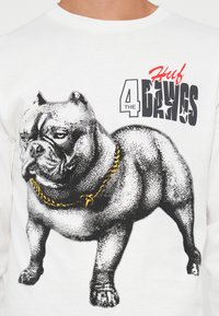 Witte sweatshirt met een afbeelding van een gespierde hond, omrand in zwart en grijs, met een gele ketting om zijn nek en vetgedrukte tekst erboven.