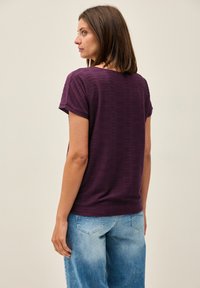 Lila randig kortärmad topp med en avslappnad passform, som har en rund halsringning och subtil textur. Kombinerad med ljusblå denimbyxor.