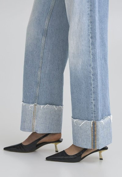 Hellblaue Denim-Jeans im weiten Schnitt mit ausgefranstem Saum und goldenen Reißverschlüssen an den Bündchen, kombiniert mit schwarzen Slingback-High Heels mit spitzen Zehen und goldenen Akzenten.
