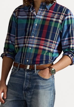 Man draagt een groen-blauw-rood geruit overhemd met opgerolde mouwen, blauwe spijkerbroek, bruine leren riem en een horloge met een groene wijzerplaat.