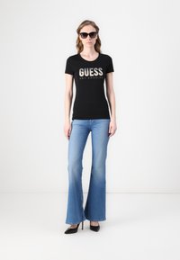 Melnas īsām piedurknēm t-kreklis ar "GUESS LOS ANGELES" metalizētām burtiem, kombinēts ar gaiši ziliem platmala džinsiem un melniem augstpapēžu apaviem.