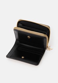 Portefeuille en cuir noir avec une fermeture éclair dorée, des emplacements pour cartes à l'intérieur et un charm en laiton avec logo. Texture lisse et forme rectangulaire compacte.