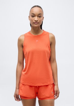 Femme debout, tournée vers l'avant, portant un haut orange sans manches et un short de sport assorti sur un fond clair uni.