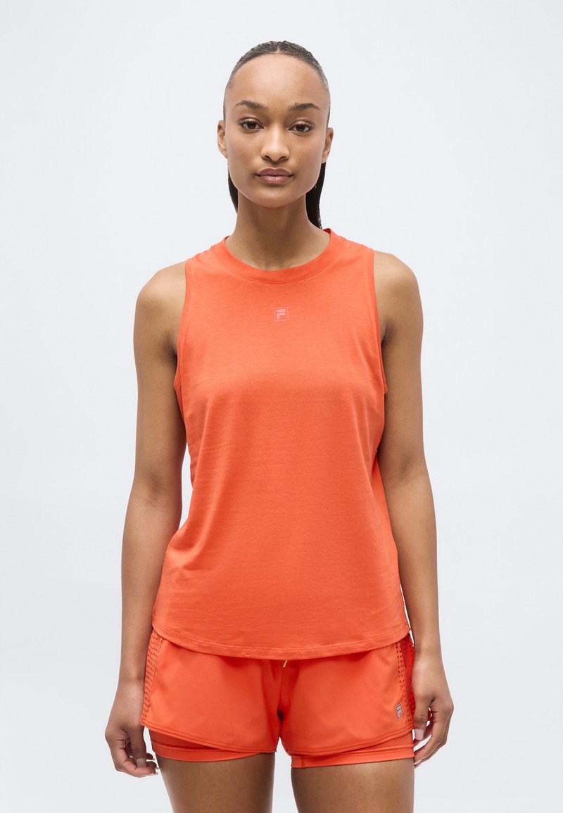 Femme debout, tournée vers l'avant, portant un haut orange sans manches et un short de sport assorti sur un fond clair uni.