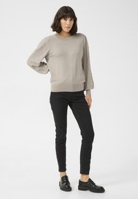 Hellgrauer Pullover mit strukturierten Ärmeln und Verzierungen, kombiniert mit schwarzen Skinny Jeans und schwarzen Slippern, vor einem schlichten Hintergrund posierend.