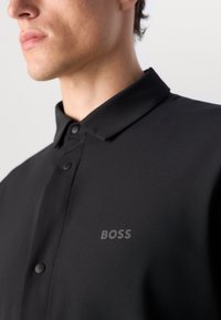 Svart polo skjorte med knapper i kragen, med en glatt tekstur og subtil "BOSS"-logo på venstre side.