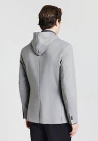 Uomo che indossa un blazer grigio chiaro con cappuccio integrato, di spalle, su uno sfondo chiaro uniforme.