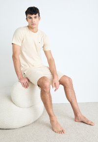 Camiseta de manga corta color crema y pantalones cortos a juego, fabricados en un tejido suave. Modelo sentada en un taburete redondeado de textura beige.
