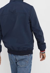 Osoba v námořnické modré bundě s pruhovanými manžetami a modrými džíny, zobrazená zezadu na jednobarevném světle šedém pozadí.