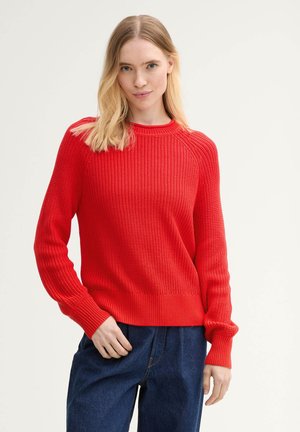 Red ribbed knit sweater with a crew neckline and long sleeves, featuring a textured pattern and fitted hem. Worn with dark blue jeans.