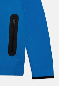 Blå jackärm med långa ärmar, med vertikal svart dragkedjeficka och svart kant vid mudd, visas mot vit bakgrund.