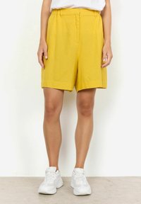 Żółte lniane szorty o luźnym kroju, z płaskim przodem i kieszeniami po bokach, w połączeniu z białymi sportowymi butami.