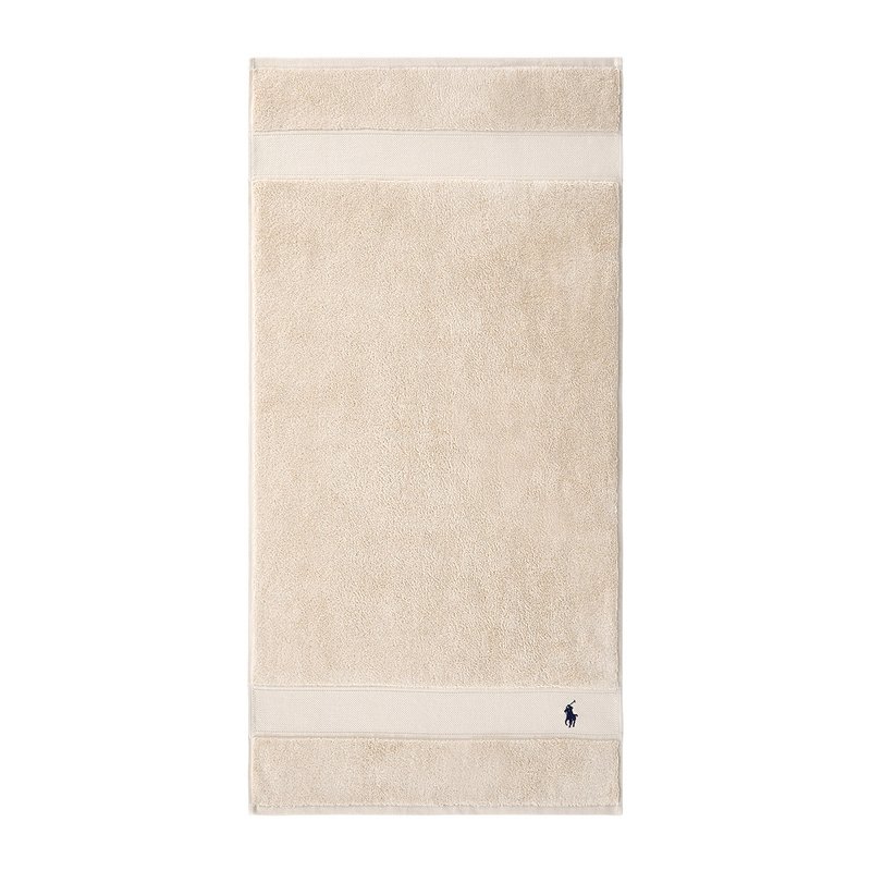 Πετσέτα μπάνιου από βαμβάκι σε μπεζ χρώμα με απα λό τερι ύφασμα, που διαθέτει μια αντίθετη κρεμ λωρίδα και ένα μικρό ναυτικό λογότυπο στην κάτω γωνία.
