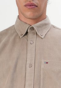 Beige corduroy shirt with a button-down collar, featuring vertical stripes, button front, and a small logo patch on the chest pocket.