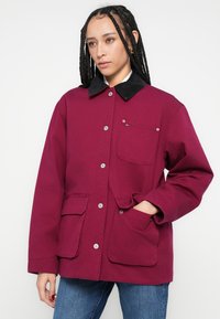 Bordowa kurtka z czarnym kołnierzem z aksamitu, srebrnymi zatrzaskami i przednimi kieszeniami. Kwadratowy fason bez wyraźnie zaznaczonej talii.