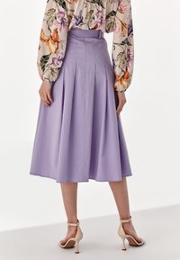 Kvinna i högmidjad lavendelfärgad midikjol, blommig långärmad blus och beigeankelremmiga högklackade sandaler, som står.