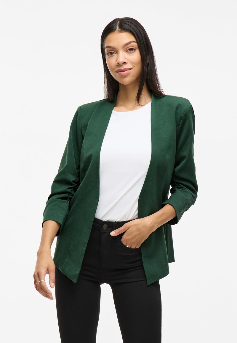 Grønn blazer med en glatt tekstur, åpen front og trekvartlange ermer, kombinert med en hvit topp og svarte bukser.