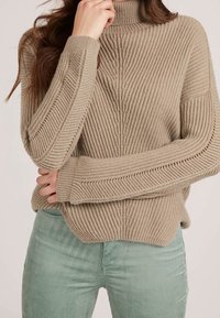 Pull en maille côtelée beige à col montant, présentant des motifs texturés verticaux et horizontaux. Associé à un pantalon en velours côtelé vert clair.