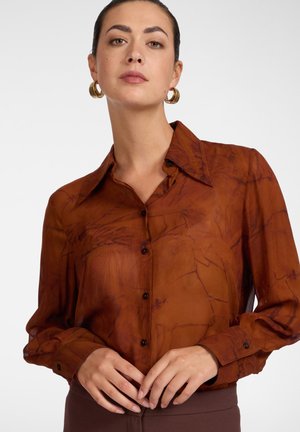 Frau mit braun gemusterter Bluse mit Knöpfen und goldenen Creolen, posiert mit locker verschränkten Händen an der Taille.