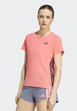 Kvinde iført koralfarvet atletisk kortærmet trøje og grå shorts med lyserøde og sorte Adidas-striber, stående mod en lys baggrund.