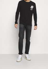 Svart långärmad T-shirt med en cirkulär planetgrafik och text på bröstet, parat med mörka jeans och vita sneakers.