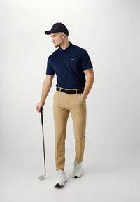 Granatowa koszulka polo z krótkimi rękawami i kołnierzykiem, khaki spodnie, czarny pasek i białe buty golfowe. Postać trzyma kij golfowy.