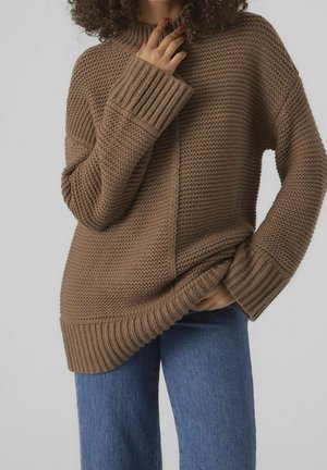 Pull en laine marron avec un col haut, des poignets côtelés et un motif texturé. Associé à un jean bleu, dont la coupe est décontractée.