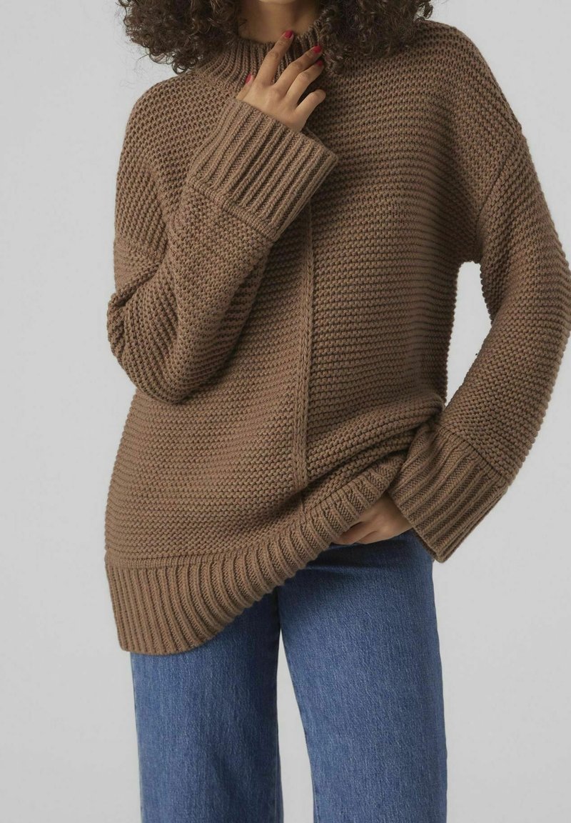 Stickad brun tröja med hög krage, ribbade ärmslut och ett strukturerat mönster. Kombinerad med blå denim, i en avslappnad passform.