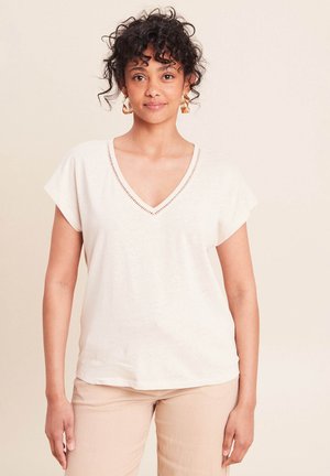 Vrouw met krullend haar, gekleed in een crèmekleurige V-hals top met korte mouwen, beige broek en gouden oorringen, staande tegen een neutrale achtergrond.