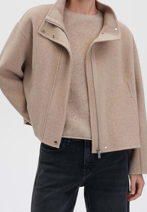 Personne portant une veste en laine beige à fermeture éclair par-dessus un pull assorti avec un jean noir, debout devant un fond clair uni.