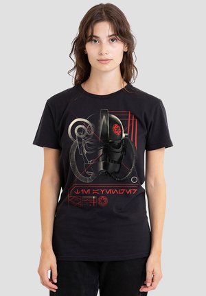 Junge Frau mit braunen Haaren, die ein schwarzes Star Wars T-Shirt trägt, das ein schwarzes Helmdesign mit roten und beigen Science-Fiction-Grafiken und Symbolen zeigt.